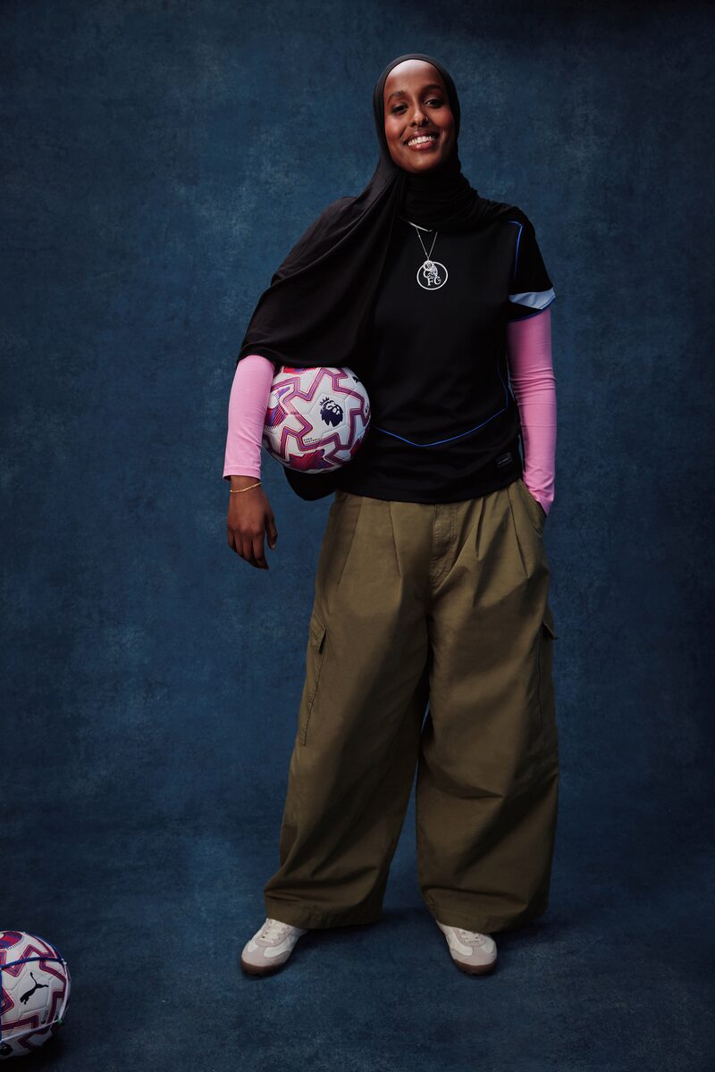Smiling woman wearing a black hijab, black Chelsea FC jersey over a pink long-sleeve top, olive cargo pants, and white sneakers, holding a Premier League football